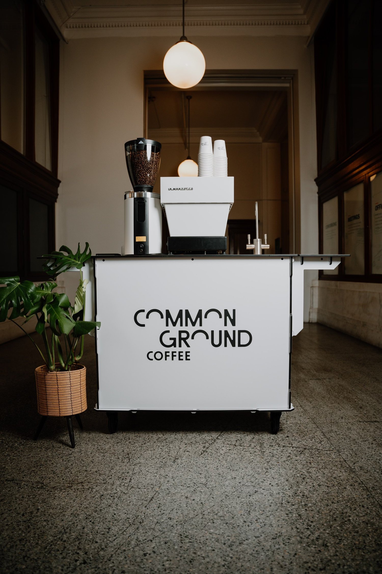 Common Ground Coffee cart with La Marzocco machine beside a monstera plant in a bright lobby